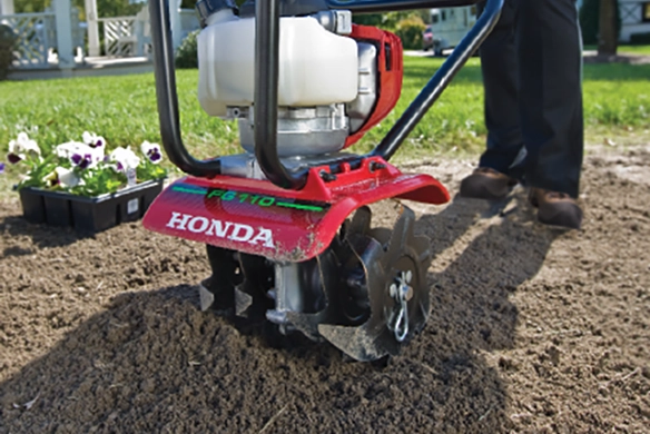 Honda FG110 mini tiller cultivating a garden bed near blooming flowers.