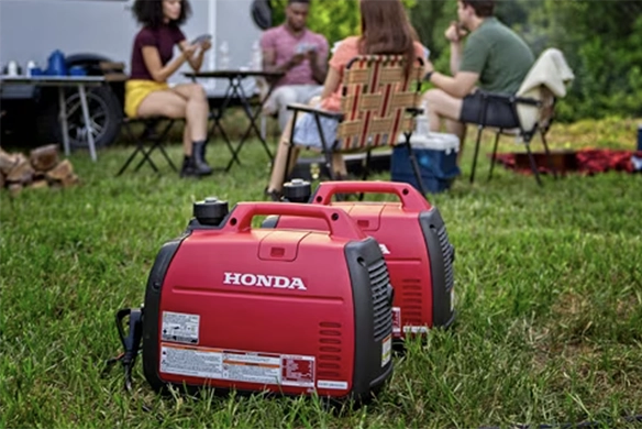 Two compact red Honda portable generators set up on grass with a group of people sitting and talking in the background.
