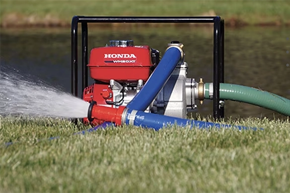 A Honda WH20X water pump actively pumping water through blue and green hoses beside a pond.