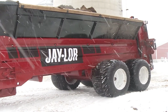 A Jaylor manure spreader trailer in snowy conditions, attached to a tractor and loaded with material.