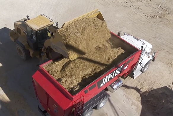 A front loader fills a Jaylor horizontal TMR feed mixer truck with silage on a dirt lot.