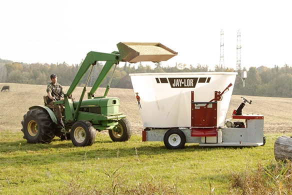 A farmer uses a tractor to load feed into a Jaylor vertical TMR mixer in an open field.