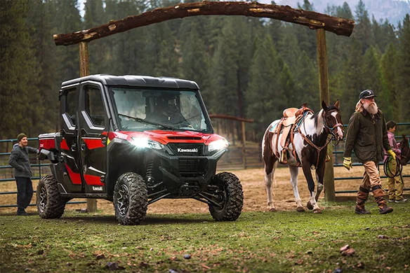 People leading a horse past a Kawasaki Ridge Crew HVAC edition in red and black, set on a ranch with rustic wooden fencing.