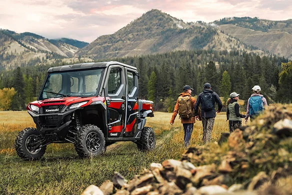 Hikers walking past a red and black Kawasaki Ridge Crew HVAC parked in a scenic mountainous meadow.