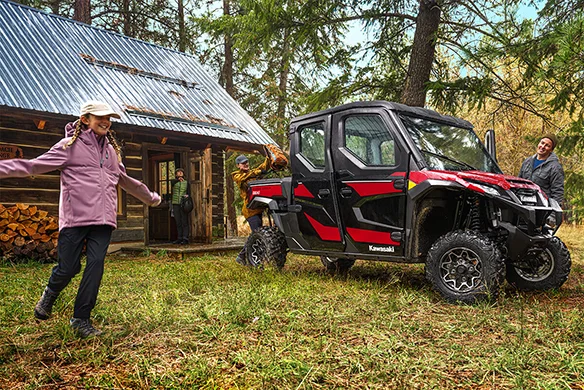 A red and black Kawasaki Ridge Crew HVAC parked outside a cozy cabin in the woods, with people gathering around playfully.