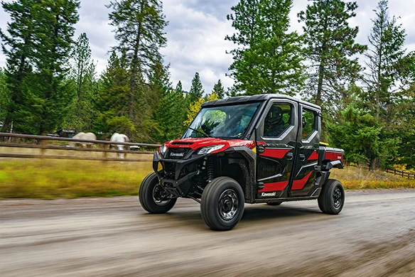 A Kawasaki Ridge Crew model in red and black passing through a ranch gate, with horses and people nearby, set in a wooded outdoor environment.