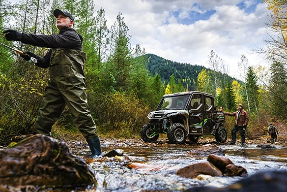 A Kawasaki Ridge XR Crew model in red and black driving quickly on a dirt road near a forest and fenced pasture.