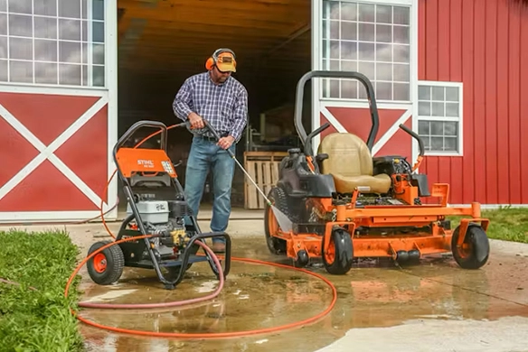 Man using a STIHL pressure washer to clean a zero-turn lawn mower in front of a barn.