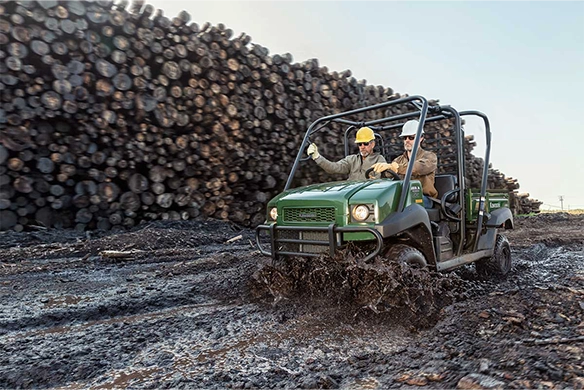 Two workers wearing hard hats drive a green Kawasaki Mule 4010 Trans 4x4 through thick mud at a logging site with stacked timber in the background.
