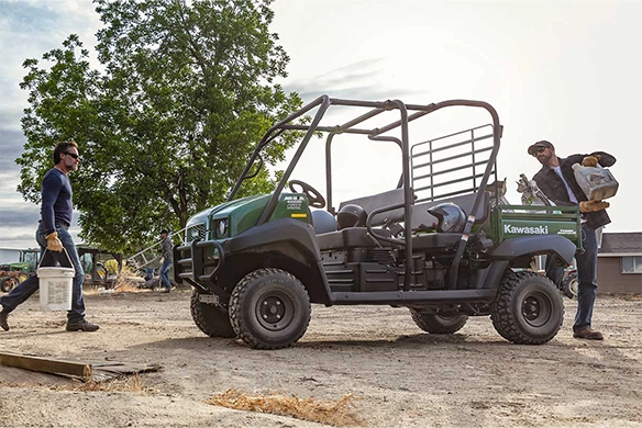 Side view of a green Kawasaki Mule 4010 Trans parked on a job site while workers carry tools and supplies.