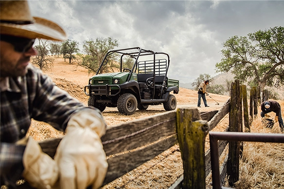 Green Kawasaki Mule 4010 Trans parked near a wooden fence while workers repair and inspect fencing in a dry, hilly field.