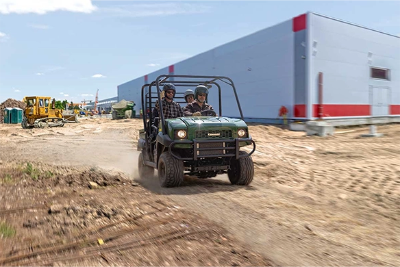 Green Kawasaki Mule 4010 Trans 4x4 driving through a construction site with three passengers onboard and heavy equipment in the background.