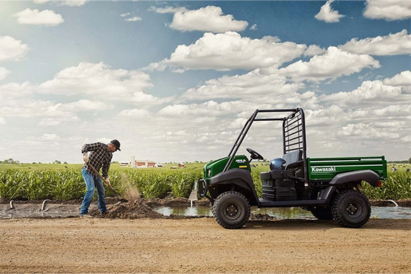 A Kawasaki Mule 4000 utility vehicle parked on a rural dirt path while a man works with a shovel near a cornfield.