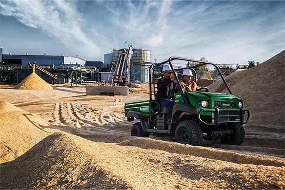 Two workers ride a green Kawasaki Mule 4000 through a dusty industrial job site with machinery in the background.