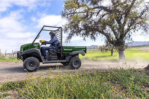 Man drives a green Kawasaki Mule 4010 down a country road with wildflowers and a large tree in view.