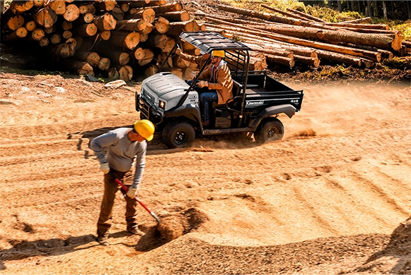 Worker operates a grey Kawasaki Mule 4010 4x4 FE while another shovels on a forestry job site surrounded by logs.