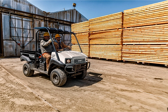 Two men in safety gear drive a grey Kawasaki Mule 4010 4x4 FE through a lumber yard.