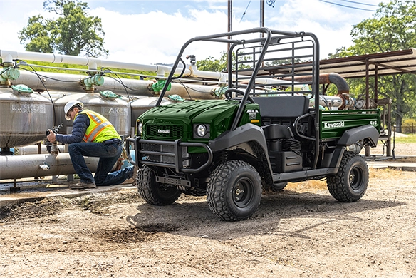 A green Kawasaki Mule 4010 utility vehicle parked near irrigation pipes while a technician performs maintenance.