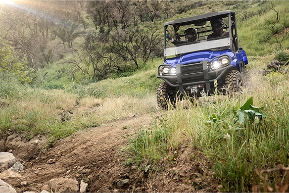 Kawasaki Mule PRO-MX LE in blue descending a rocky trail with two passengers navigating through a grassy hillside.