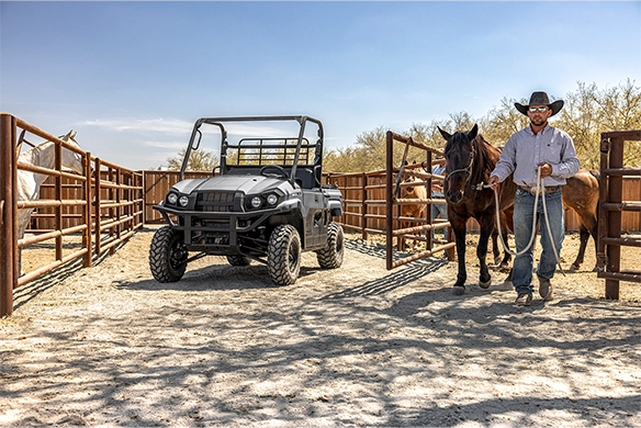 A black Kawasaki Mule PRO-MX parked in a livestock corral as a rancher leads two horses nearby.