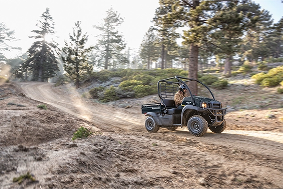 A green Kawasaki Mule SX speeds down a winding trail in a forested area, driven by a single rider.
