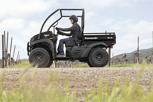 A rider drives a black Kawasaki Mule SX 4x4 through a vineyard on a dirt path, with hills visible in the background under a bright sky.