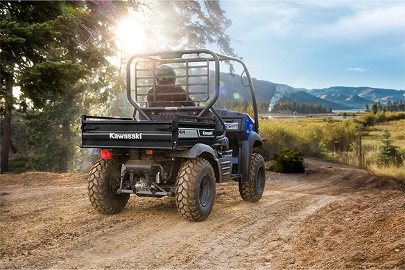 Rear view of a blue Kawasaki Mule SX 4x4 XC driving on a dirt trail through scenic countryside at sunset.