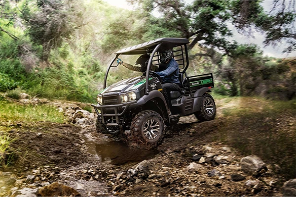 A rider navigates a rocky off-road path in a black Kawasaki Mule SX 4x4 XC LE, splashing through muddy terrain.