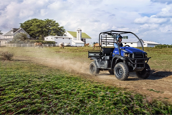Blue Kawasaki Mule SX 4x4 XC speeds past a farm with horses in the pasture and barns in the background.