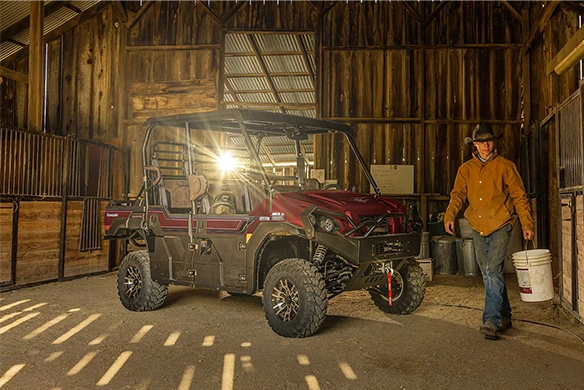 A red Kawasaki Mule PRO-FXT 1000 parked inside a barn as a rancher walks by with a bucket, sunlight streaming through wooden slats.