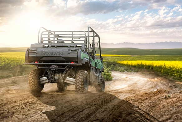 Rear view of a Kawasaki Mule PRO-FXT 820 kicking up dust on a winding dirt road surrounded by yellow fields and rolling hills at sunrise