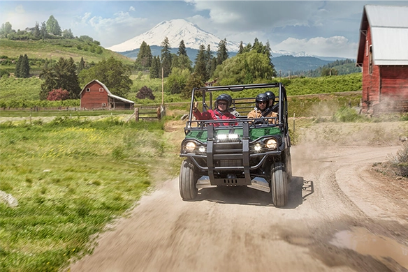 Three passengers ride a Kawasaki Mule PRO-FXT 820 through a scenic farm road, with red barns and snow-capped mountains in the background.