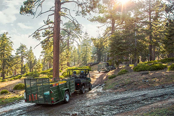Two Kawasaki Mule PRO-DX utility vehicles haul trailers along a wooded trail with sun filtering through the trees.