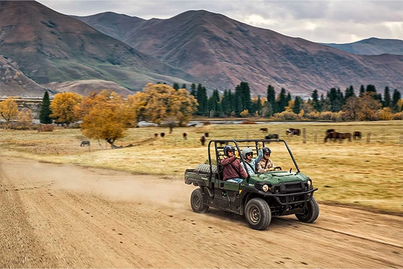 Green Kawasaki Mule PRO-DX kicks up dust as it drives through a ranch with cattle grazing and mountains in the background.