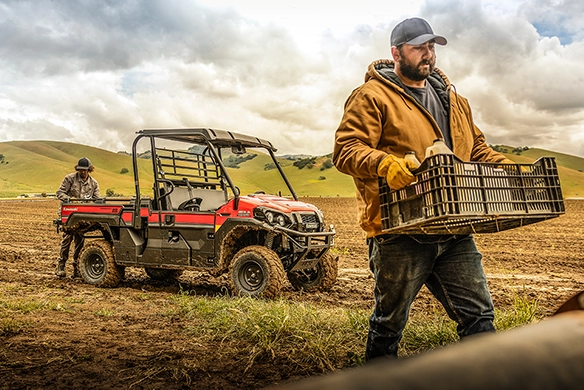 Workers transport crates in a field using a red Kawasaki Mule PRO-FX1000 HD utility vehicle on a cloudy day.