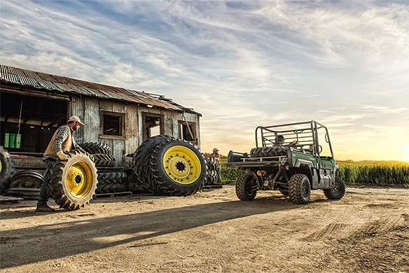 A Kawasaki Mule PRO-FX 820 is parked near an old farm building while workers roll large tractor tires at sunset.