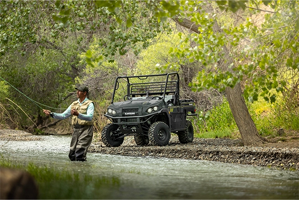 A man fly fishing in a stream while a black Kawasaki Mule PRO-FXR 1000 is parked on the gravel bank under dense tree cover.