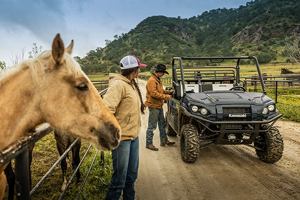 Two ranchers talk near a black Kawasaki Mule PRO-FXR 1000 as horses stand by the fence and rolling hills rise in the background.