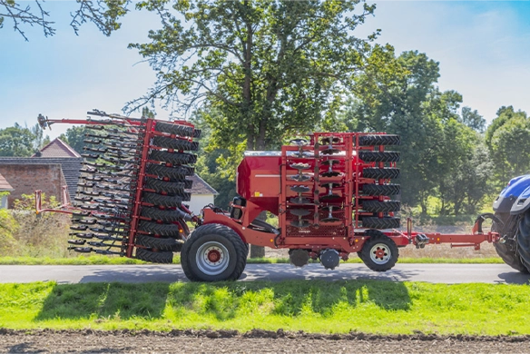 Horsch tillage implement folded for transport, pulled by a tractor on a rural road with trees in the background.