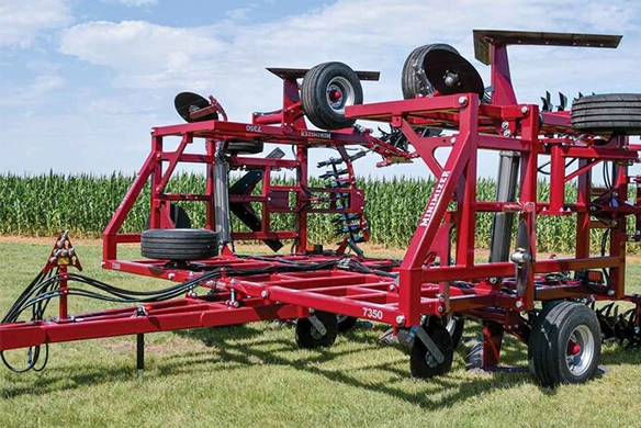 Red Unverferth tillage implement displayed on grass in front of a cornfield under a blue sky.