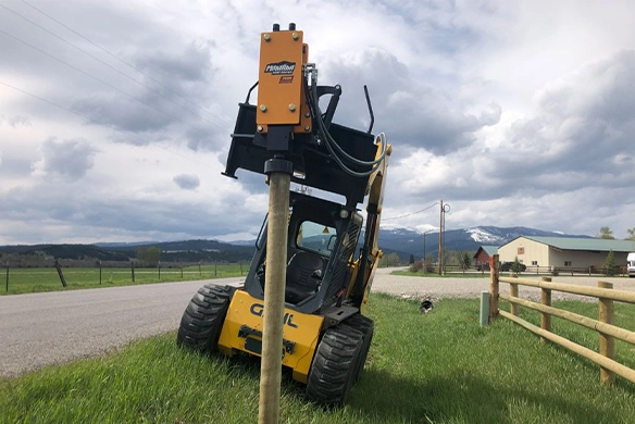 Montana Post Driver 750R mounted on a skid steer, driving a post beside a country road.