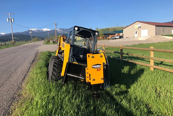 Montana Post Driver 750R attached to a skid steer, parked near a fence line in the grass.
