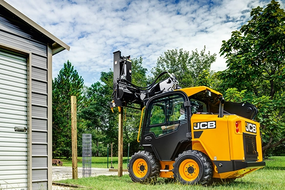 JCB 215 skid steer loader fitted with post driver attachment installing fence posts beside a building