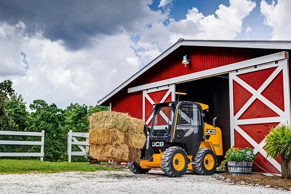 JCB 300 skid steer loader carrying stacked hay bales outside a farm barn