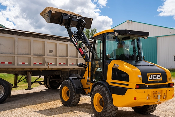 JCB 407 wheel loader dumping material from its raised bucket into a flatbed trailer at an outdoor work site