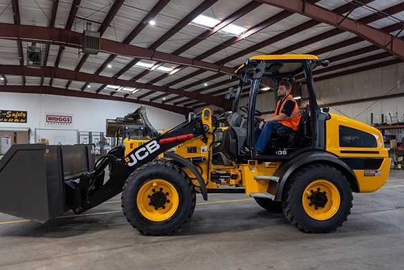 Operator driving JCB 409 wheel loader inside an indoor warehouse facility with high ceiling and steel beam construction