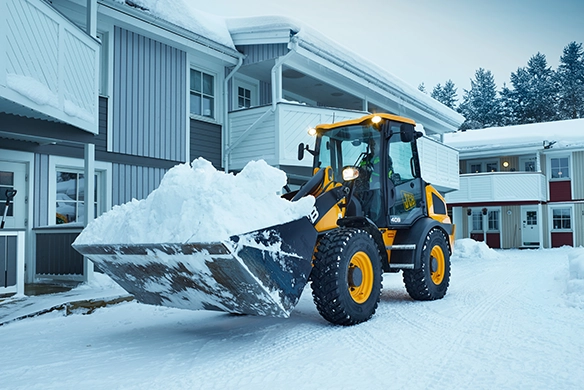 JCB 409 wheel loader clearing snow with a full bucket in front of a residential building during winter