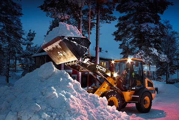 JCB 409 wheel loader dumping snow at dusk with illuminated cab lights in a snowy residential area