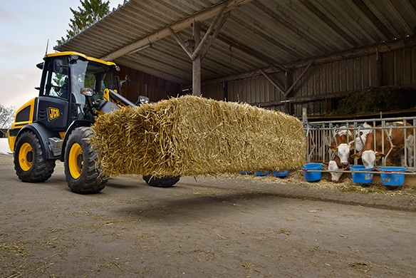 JCB 409 wheel loader transporting a large hay bale with hay spike attachment in a covered barn with cattle visible in the background