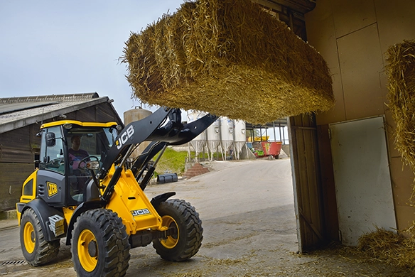 JCB 409 wheel loader lifting a large hay bale with front-mounted bale spear at a farm storage facility